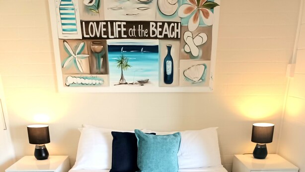 A neatly made bed with a teal throw blanket and two pillows, flanked by white nightstands with lamps, in a room with a beach-themed wall art. - Golden Beach Holiday Park (Golden Beach)