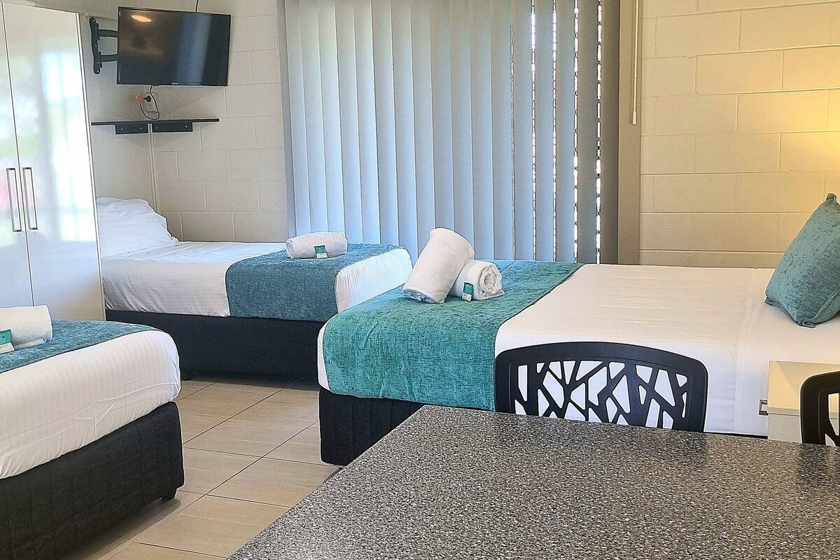 Habitación de hotel con dos camas, un televisor montado en la pared, una mesa con sillas y una ventana con persianas.