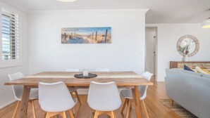 A dining area with a wooden table, white chairs, a painting of a beach, and a round mirror on the wall.