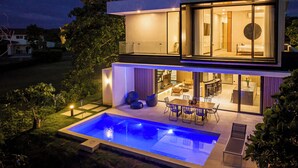 Ett modernt hus i två våningar med en swimmingpool, en uteplats och en stor glasdörr som leder in till huset.