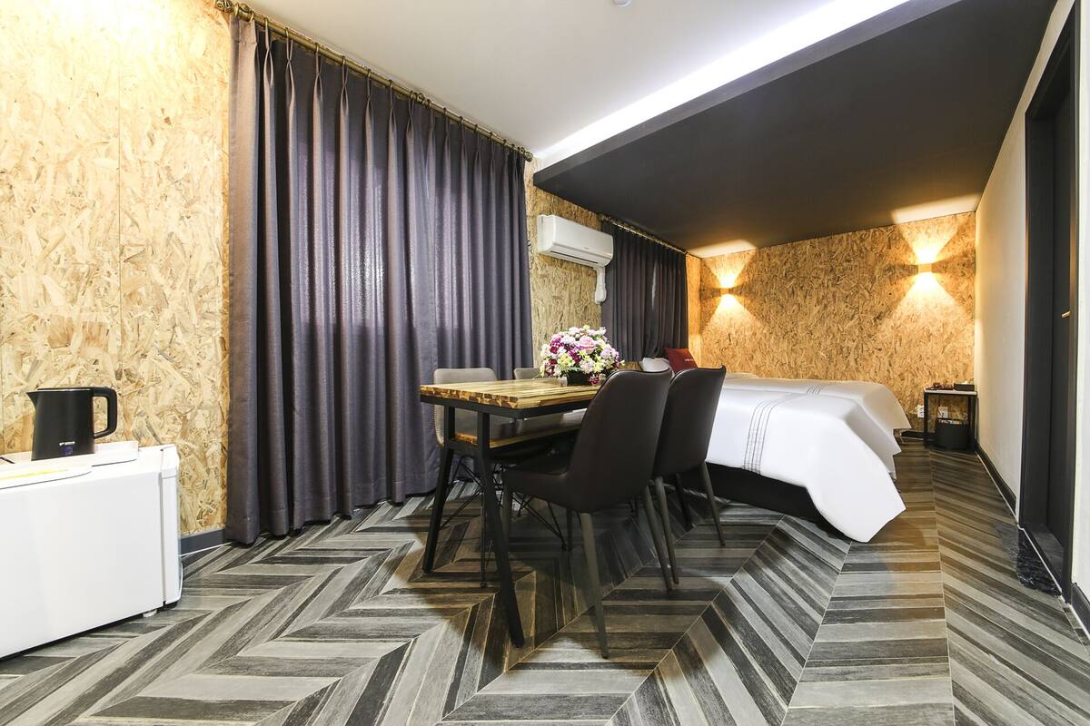 Ein Hotelzimmer mit Fischgrätparkett, einem Holzespeisetisch mit Stühlen, einem Bett mit weißer Bettdecke und einer Wand-Klimaanlage.