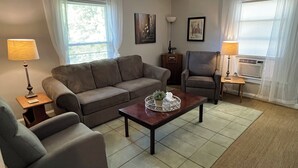 Una sala de estar con sofá, sillón y mesa de centro. Hay dos lámparas, un marco de fotos y una pequeña mesa con una planta.