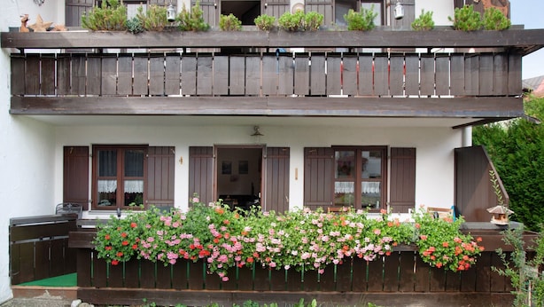 Edifici de dues plantes amb un balcó, persianes de fusta i un jardí de flors al davant.