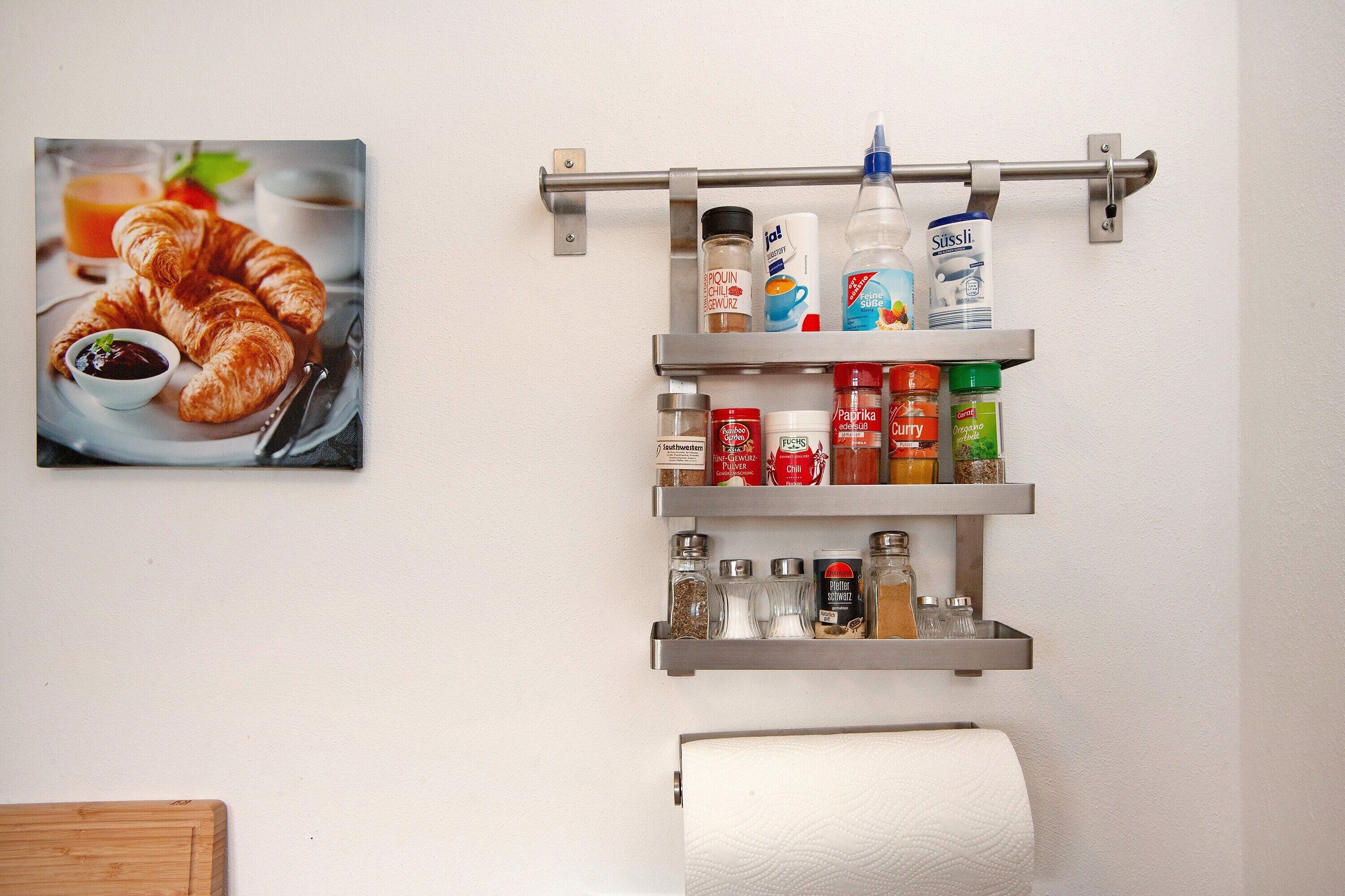 Een keuken met een hangplank vol kruiden en specerijen, een afbeelding van croissants, en een rol keukenpapier.