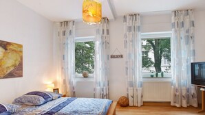 Un dormitorio con una cama, un televisor, un ventanal con cortinas, un radiador y una pared con un cartel que dice 'BELIEVE'.