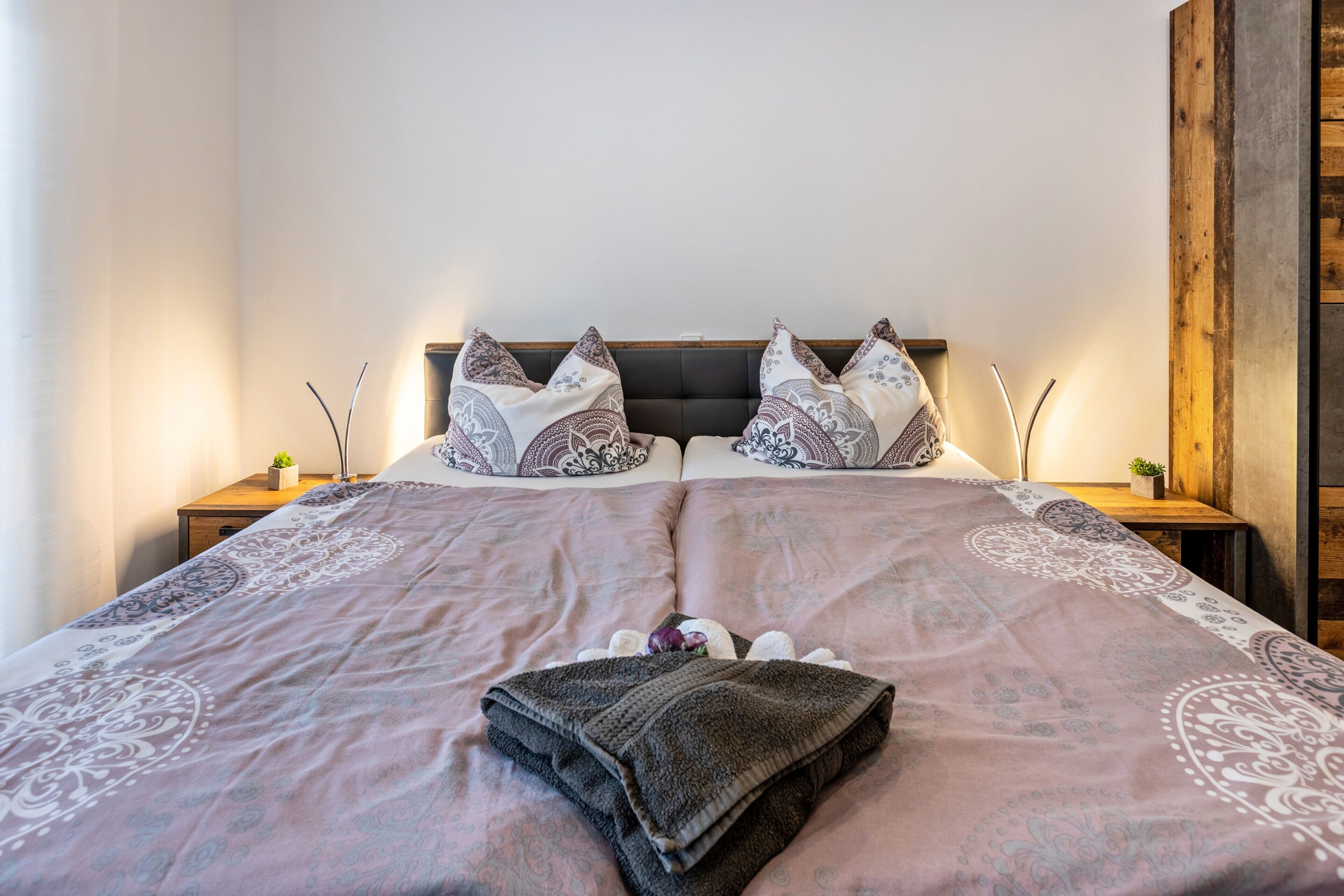 A neatly made bed with patterned bedding, two bedside tables with small plants, and a wooden headboard.