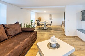 Een moderne woonkamer met een bruine leren bank, een witte salontafel met kaarsen, een eethoek met tafel en stoelen, en een groot raam dat natuurlijk licht binnenlaat.