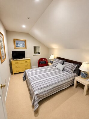A bedroom with a bed, a television on a dresser, a chair, and a lamp on a bedside table.
