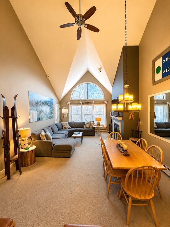 A spacious living area with a high ceiling fan, a dining table with chairs, a fireplace, and a large window with a view.
