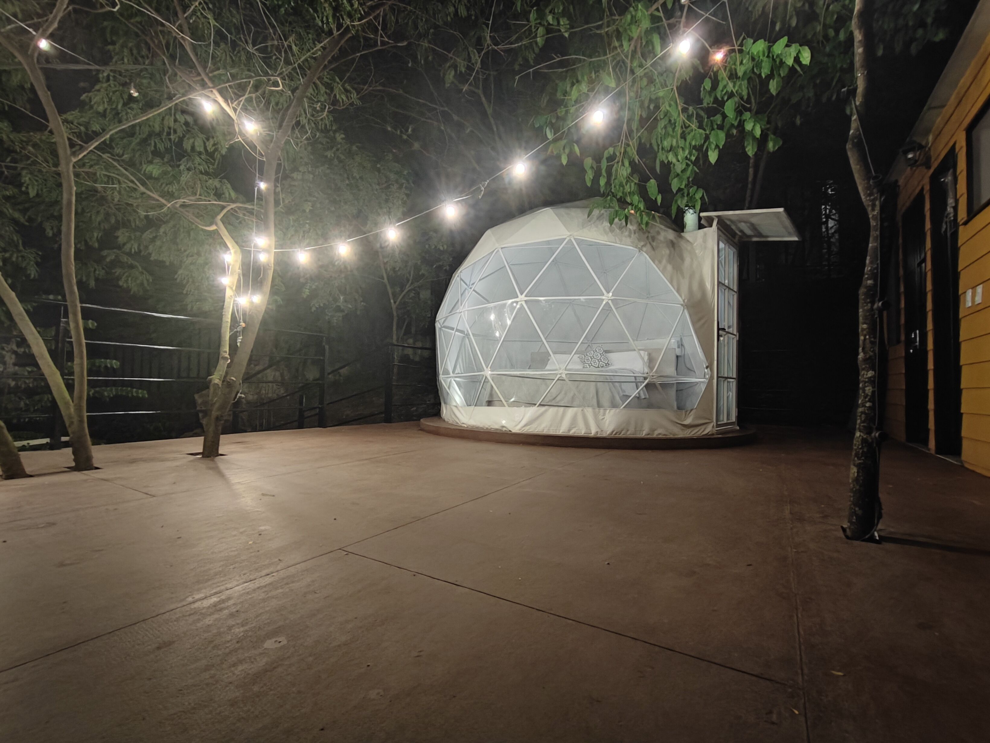 Uma tenda em forma de cúpula geodésica com uma cama no interior, rodeada por árvores e luzes de corda.