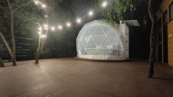 Uma tenda em forma de cúpula geodésica com uma cama no interior, rodeada por árvores e luzes de corda.