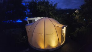 Une structure en forme de dôme, ornée d’une guirlande lumineuse, entourée d’arbres et avec une montagne en arrière-plan.