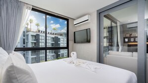 A hotel room with a large bed, a flat-screen TV, an air conditioning unit, and a view of a building with palm trees. - Citygate Kamala Residence P506  (Kamala)