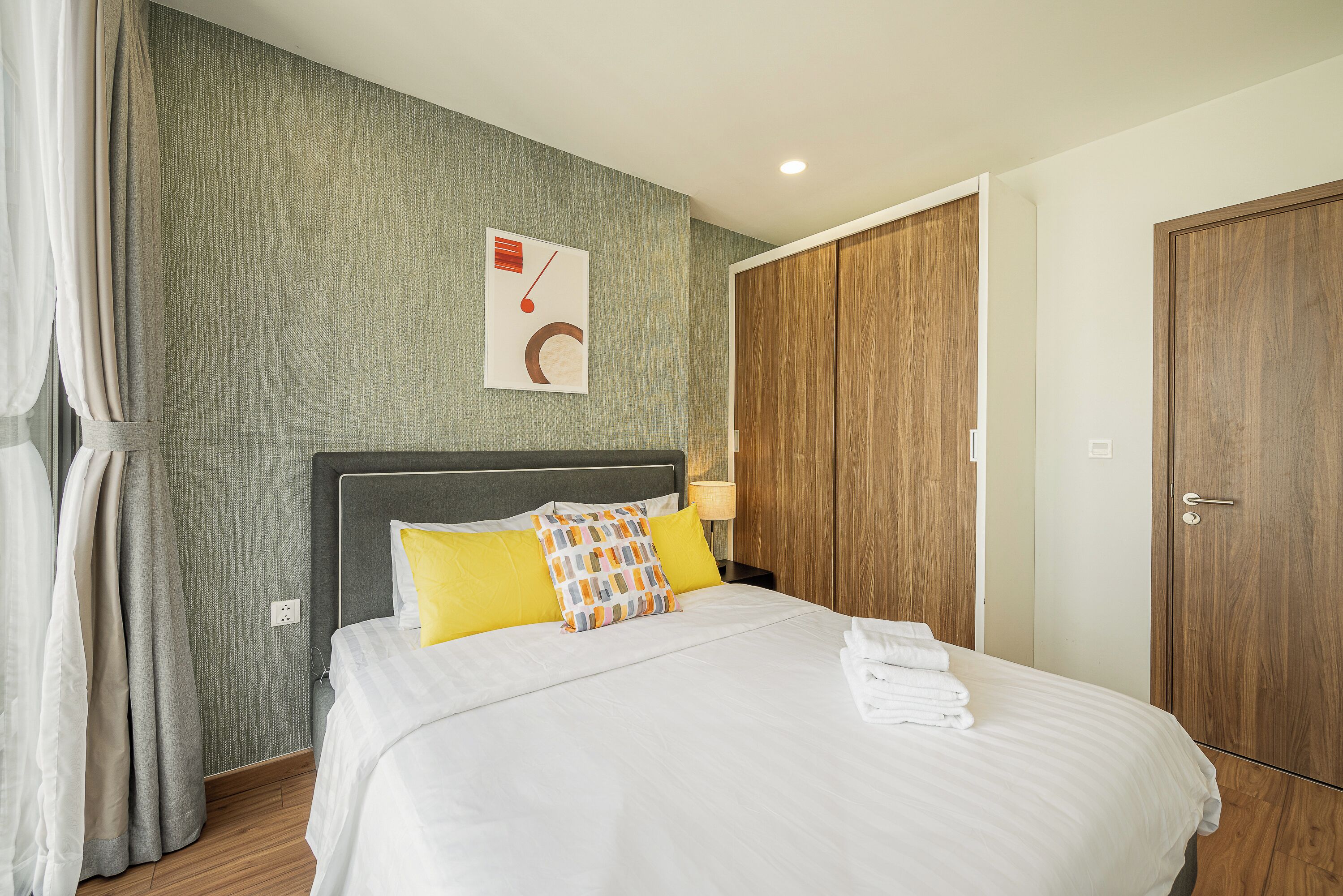 A neatly made bed with white linens and a colorful pillow, a wooden wardrobe, and a wall-mounted artwork.
