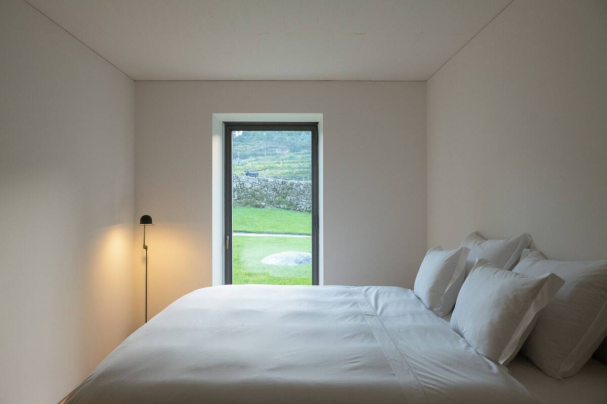 Une chambre à coucher avec un lit, une table de chevet et une lampe. Il y a une fenêtre donnant sur un jardin.