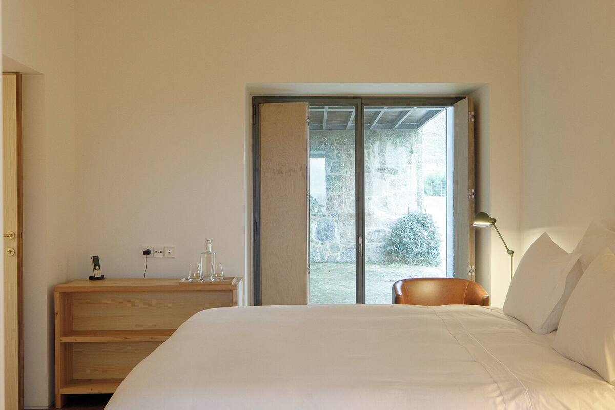 Une chambre à coucher au style minimaliste, avec un grand lit, une table de chevet en bois, un bureau et une vue sur l’extérieur grâce à une porte vitrée.