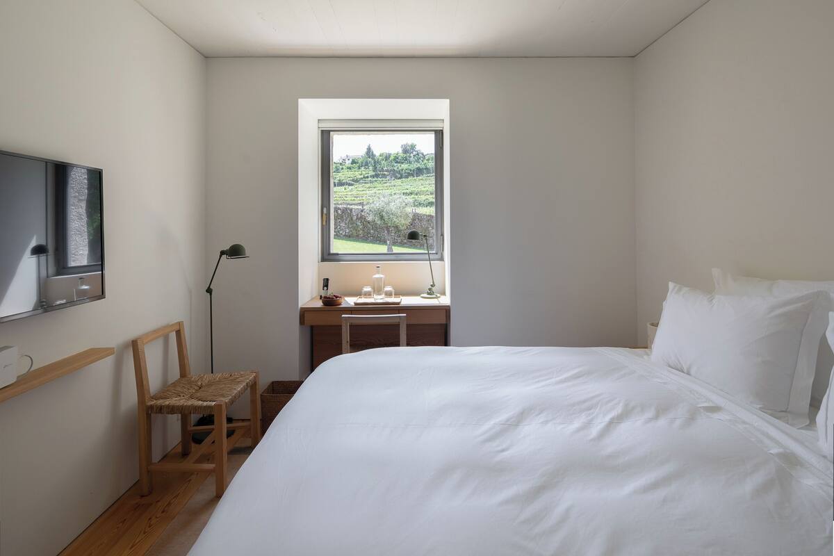 Une chambre avec un lit, un bureau et une chaise, un téléviseur fixé au mur et une fenêtre donnant sur un paysage verdoyant.