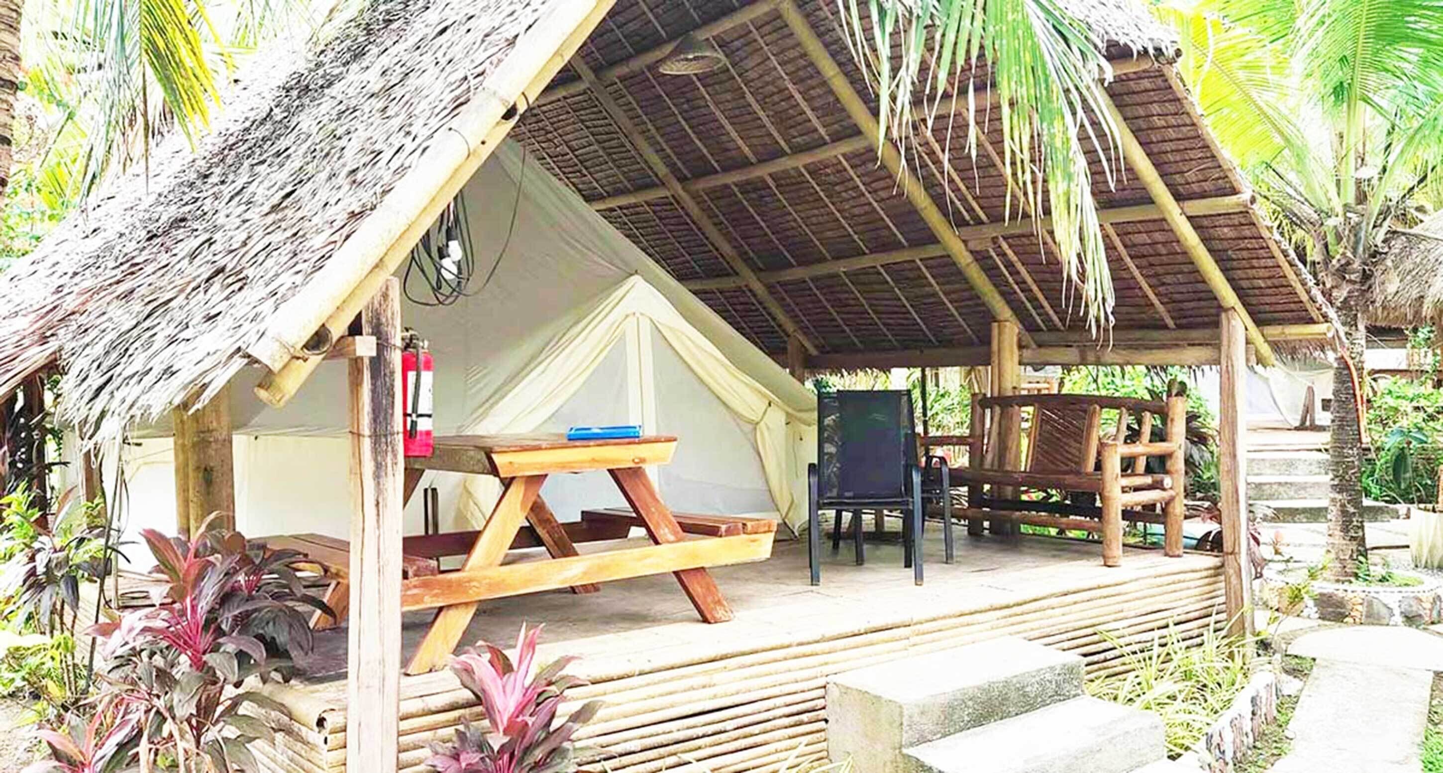 A thatched-roof bungalow with a wooden deck, a table, and chairs.