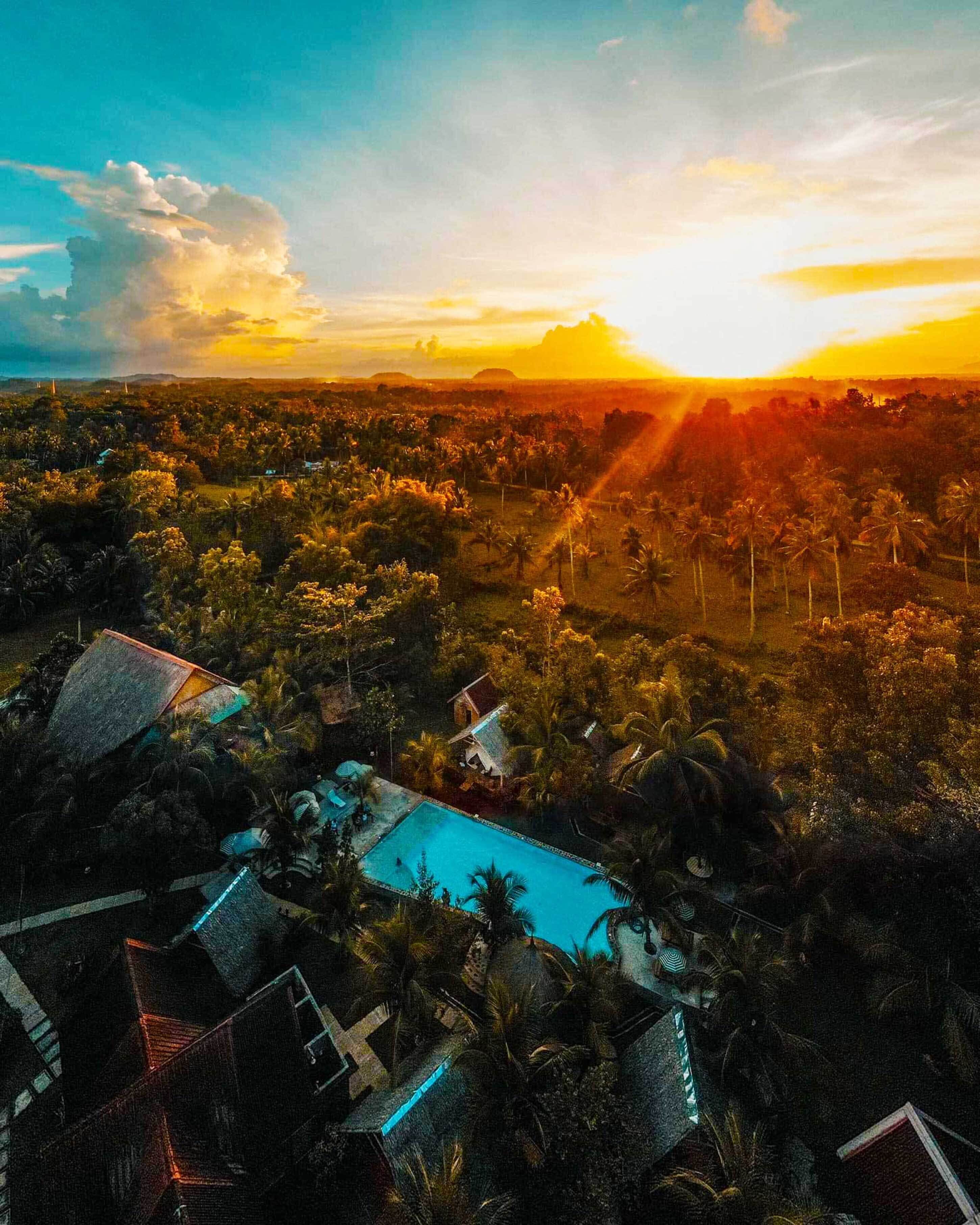 A resort with multiple buildings, a swimming pool, and a view of the sunset over a landscape with palm trees.