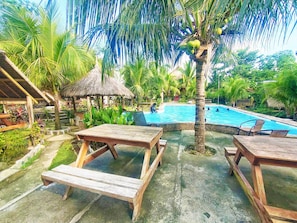 A pool area with wooden tables and benches, surrounded by lush greenery and palm trees.