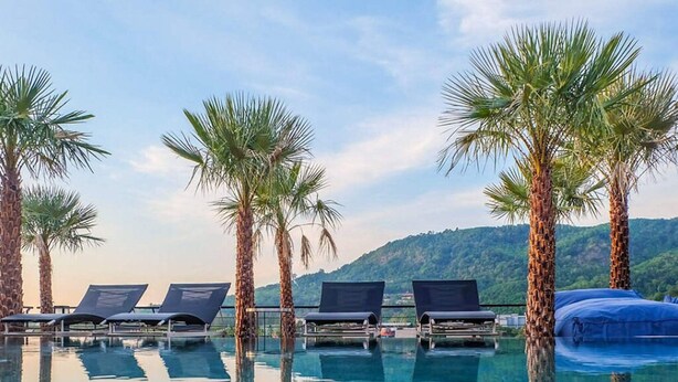 A poolside area with lounge chairs and palm trees, surrounded by a mountainous landscape. - Citygate Kamala Residence P204 (Kamala)