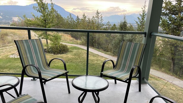 A balcony with four striped chairs and a small table, overlooking a grassy area with trees and mountains in the background.