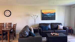 A living room with a dining table, leather sofas, a coffee table, a clock, a floor lamp, and a painting of a mountain landscape.