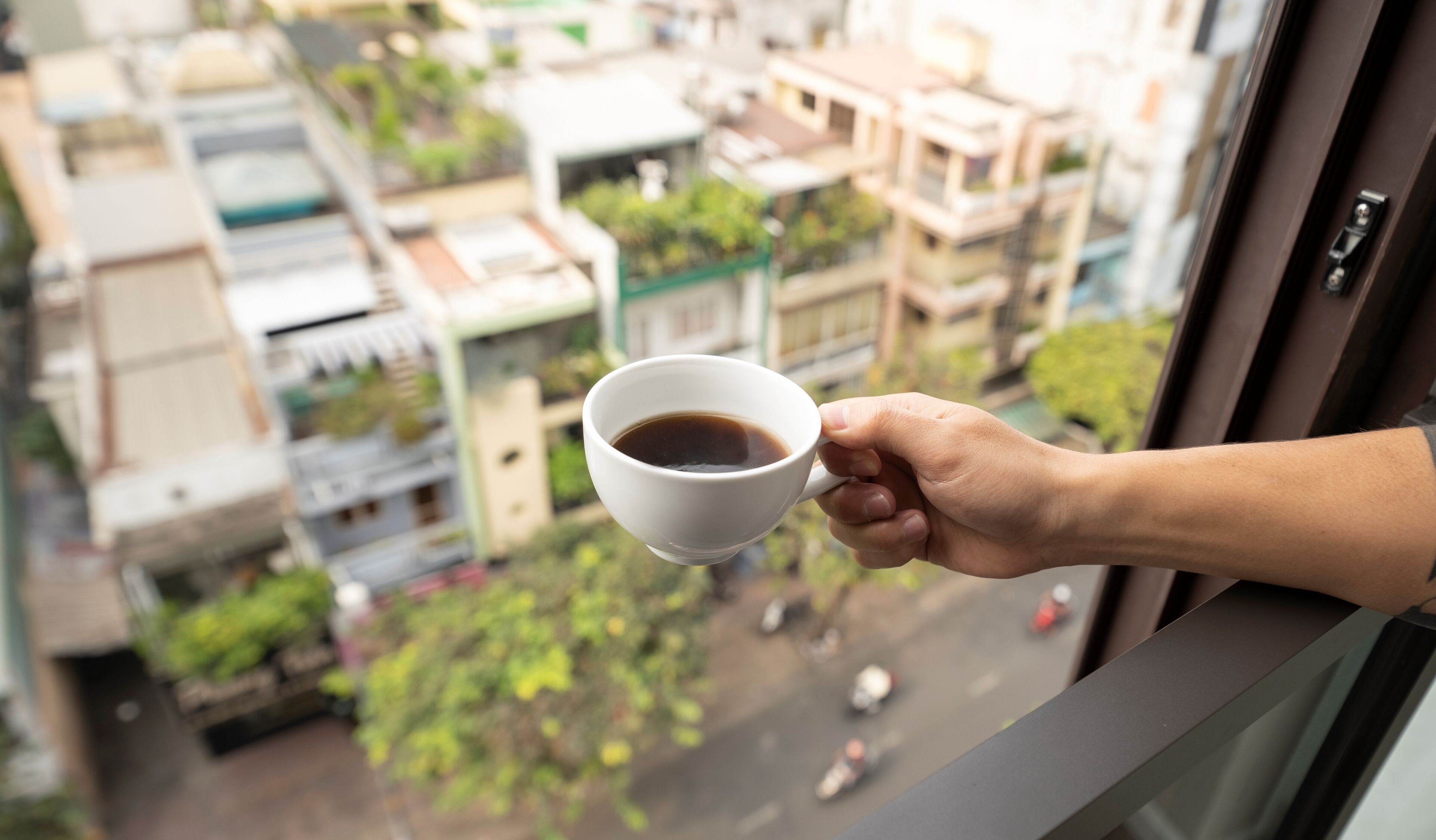 A hand holding a white cup of coffee, with a view of a residential area through a window.