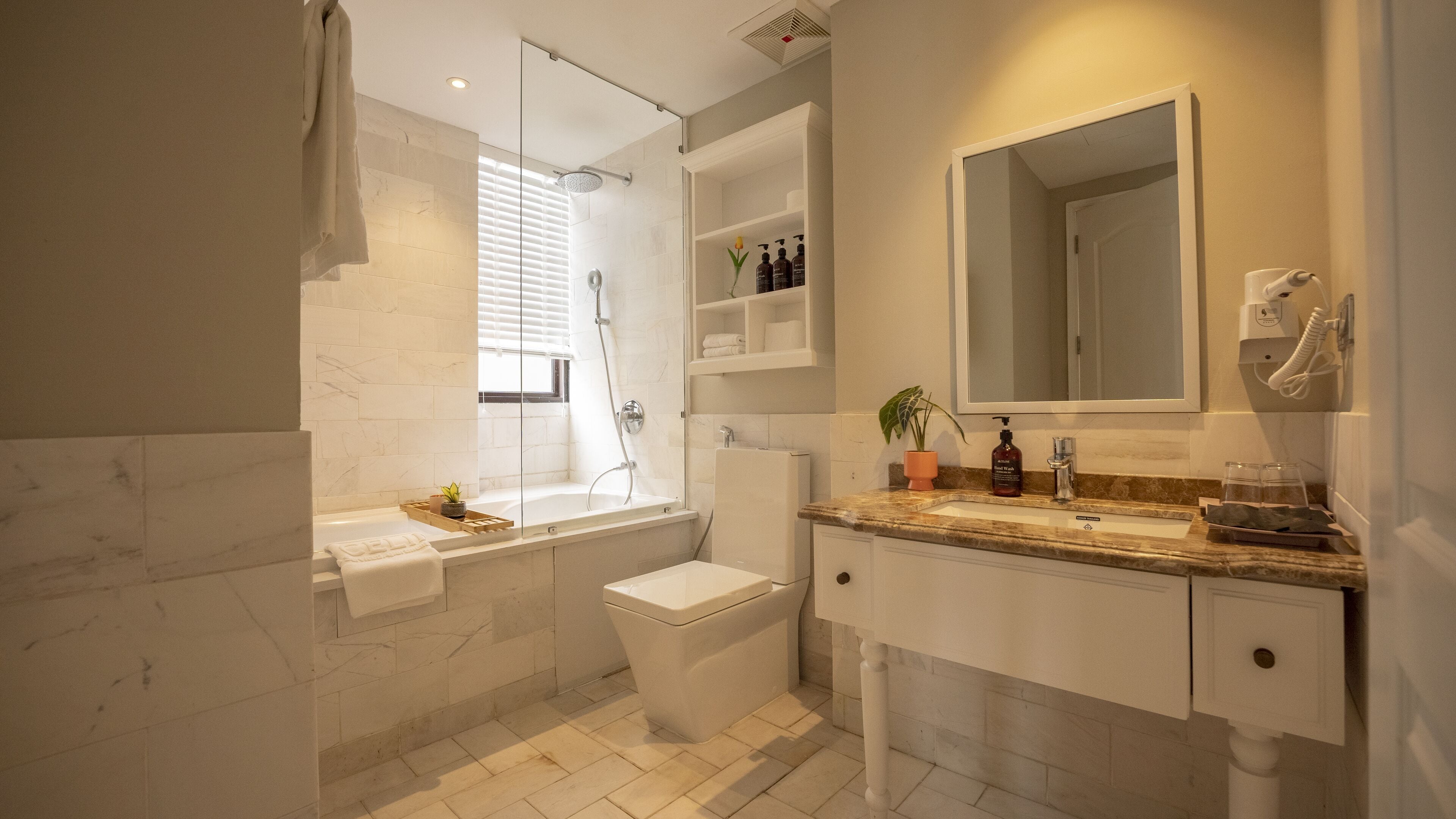 A modern bathroom with a bathtub, toilet, and sink. There is a window with blinds, a mirror, and a shelf with toiletries.