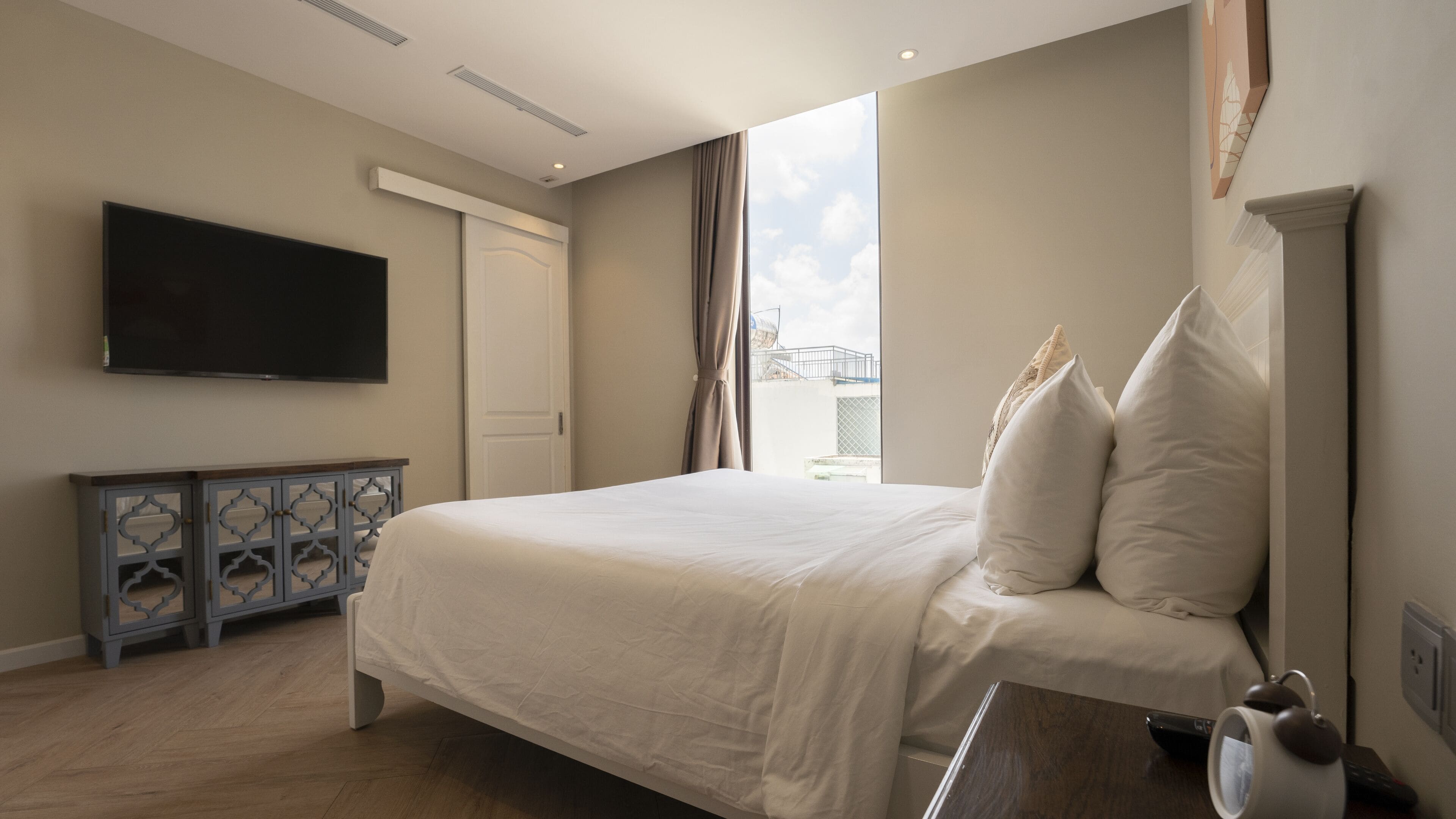 A bedroom with a bed, a TV mounted on the wall, a window with curtains, and a small table with a kettle.
