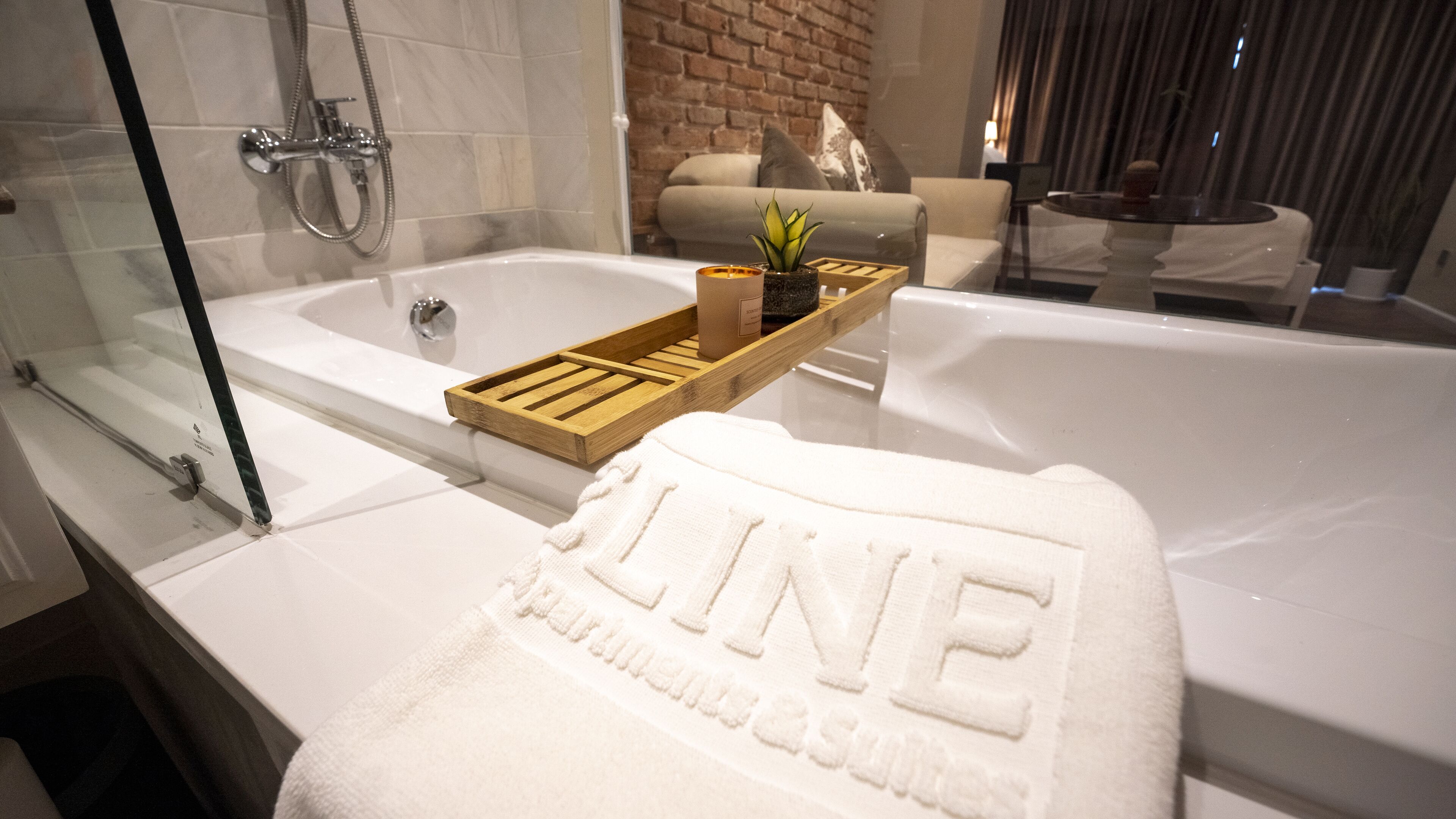 Un baño moderno con ducha de vidrio, una bandeja de madera con artículos de aseo y una toalla con la palabra 'LINE' bordada.