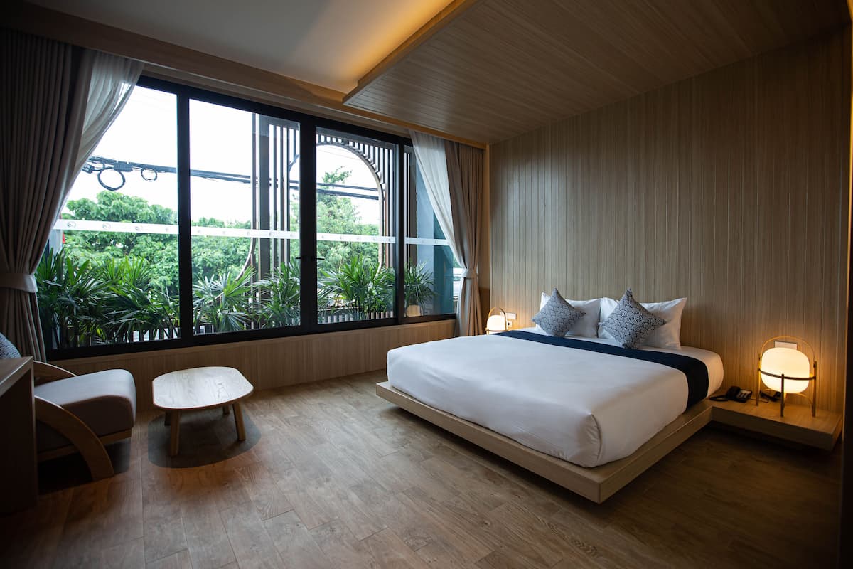A modern hotel room with a large bed, a small seating area, and a view of greenery through the window.