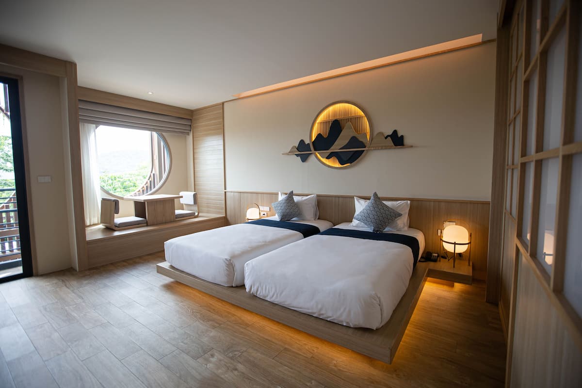 A modern hotel room with two beds, a desk, and a view of greenery.