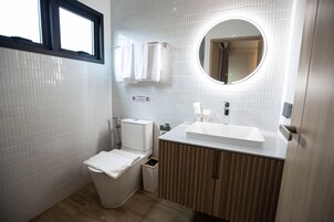 Un baño moderno con paredes de azulejos blancos, un tocador de madera, un espejo redondo y un inodoro con una toalla sobre el asiento.