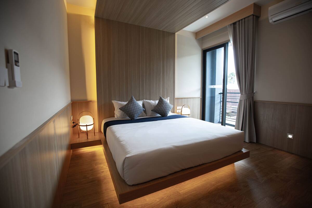 A modern hotel room with a large bed, wooden flooring, a built-in bench, a bedside table with a lamp, and a view of the outdoors through a large window.