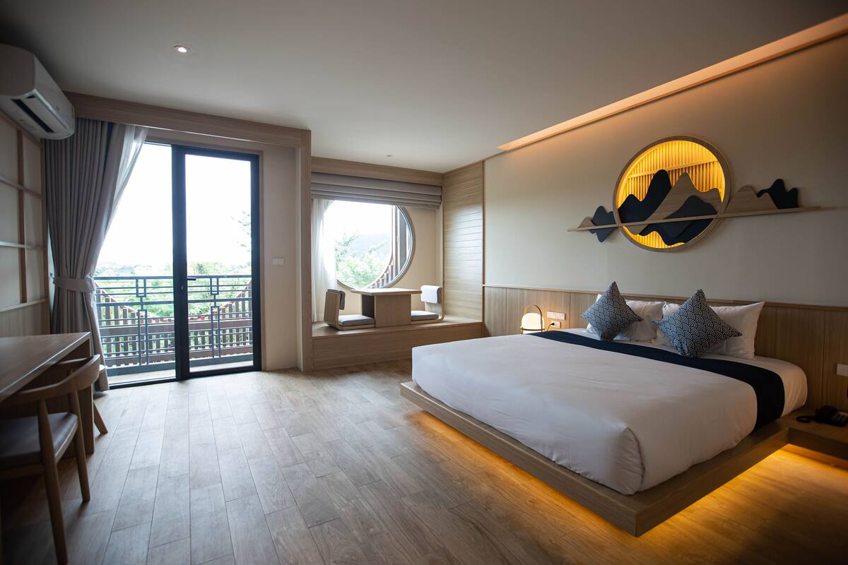 A hotel room with a large bed, a desk, a chair, a window with a view, and a wall-mounted artwork.