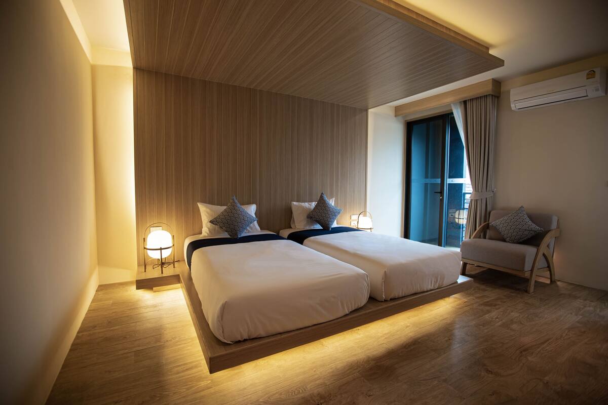 A modern hotel room with two beds, a wooden ceiling, a window with curtains, and an air conditioning unit.