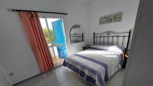 Un dormitorio con cama, una ventana con cortinas, un espejo fijado a la pared y una mesita de noche pequeña.