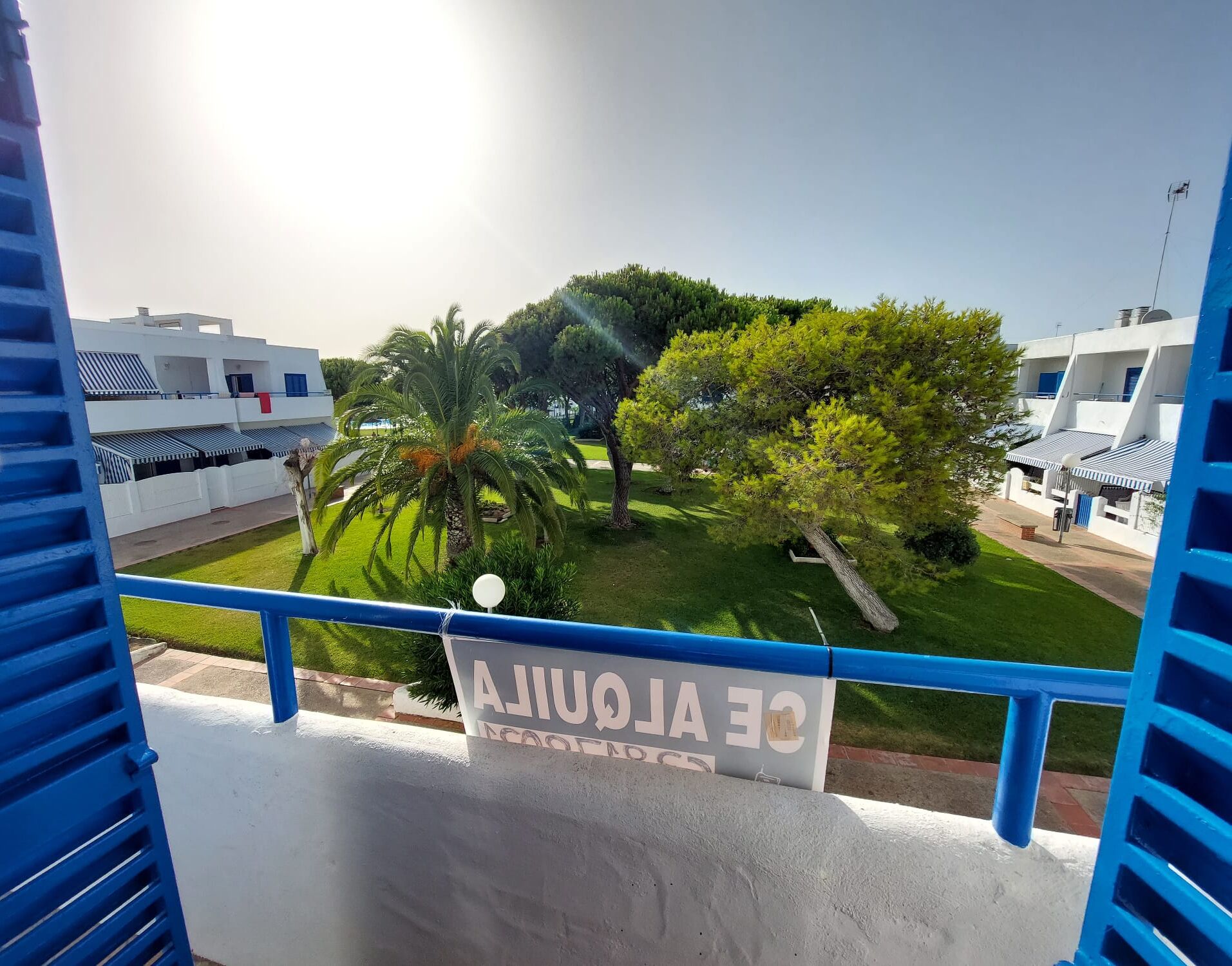 Een balkon met uitzicht op een verzorgde tuin, een palmboom en een bord met de tekst 'CE ALQUILA'.