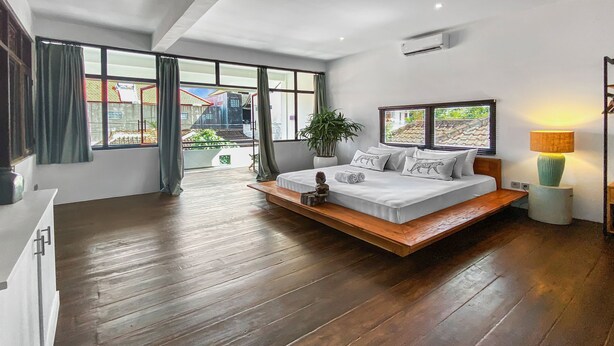 A modern bedroom with a wooden floor, a bed with white bedding, a bedside table, a lamp, and a view of the outside through large windows. - Berawa Factory Upstairs (Canggu)