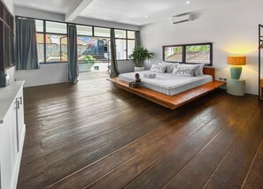A modern bedroom with a wooden floor, a bed with white bedding, a bedside table, a lamp, and a view of the outside through large windows. - Berawa Factory Upstairs (Canggu)
