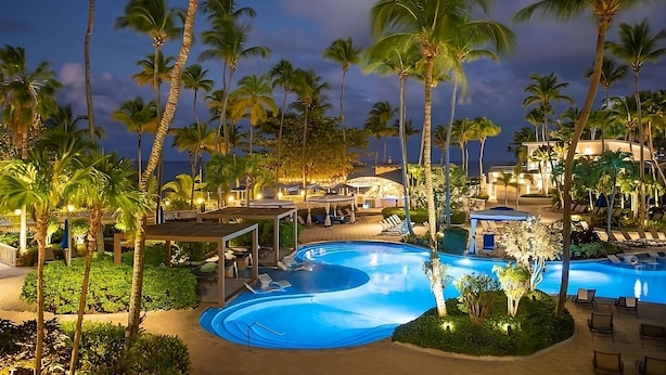 Ein Poolbereich eines Resorts mit Palmen, Liegestühlen und einer überdachten Sitzgelegenheit im Abendlicht.
