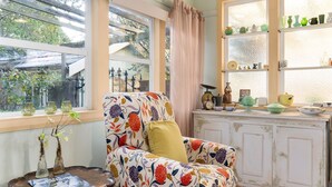 A cozy room with a floral-patterned armchair, a window with curtains, and a shelf with various decorative items. - Secret Garden Cottage (Katoomba)