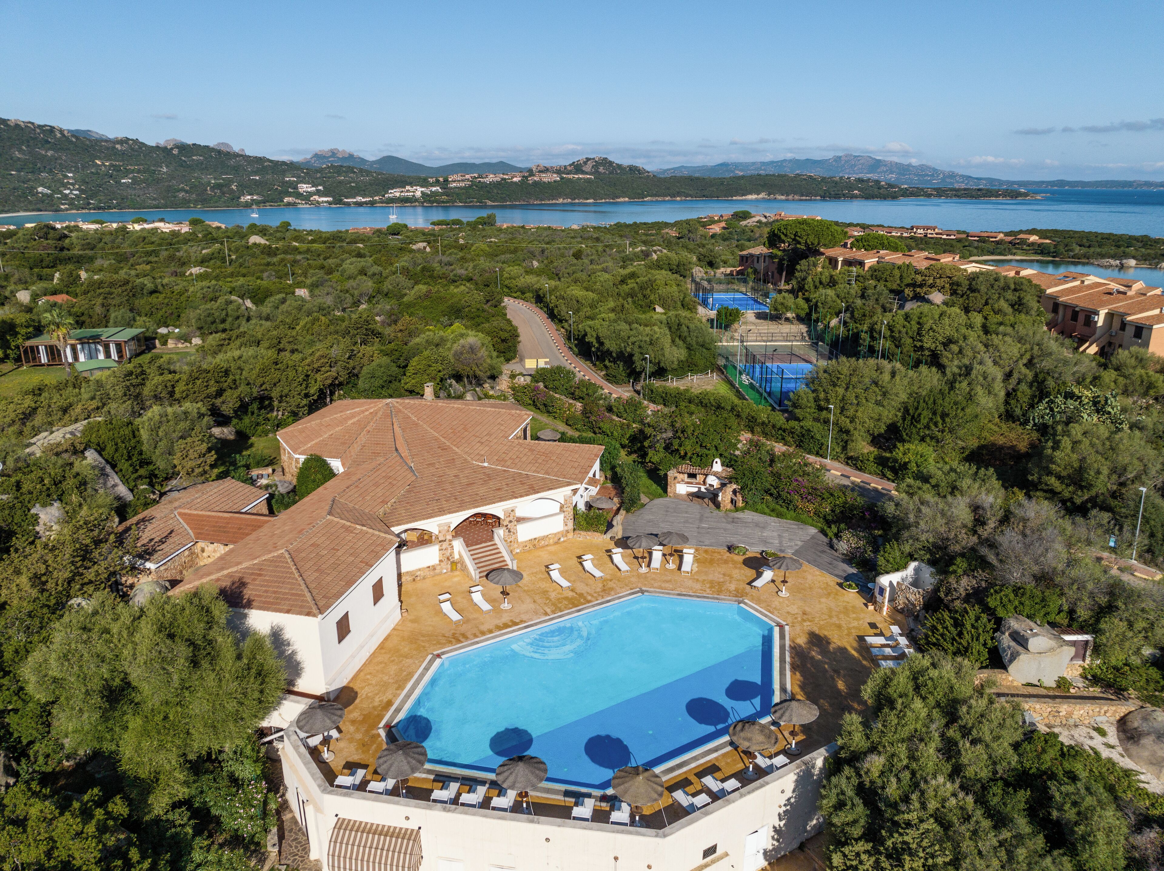 Vista aerea di un resort con piscina circondata da lettini e ombrelloni, con sullo sfondo un paesaggio costiero, montagne e cielo sereno.