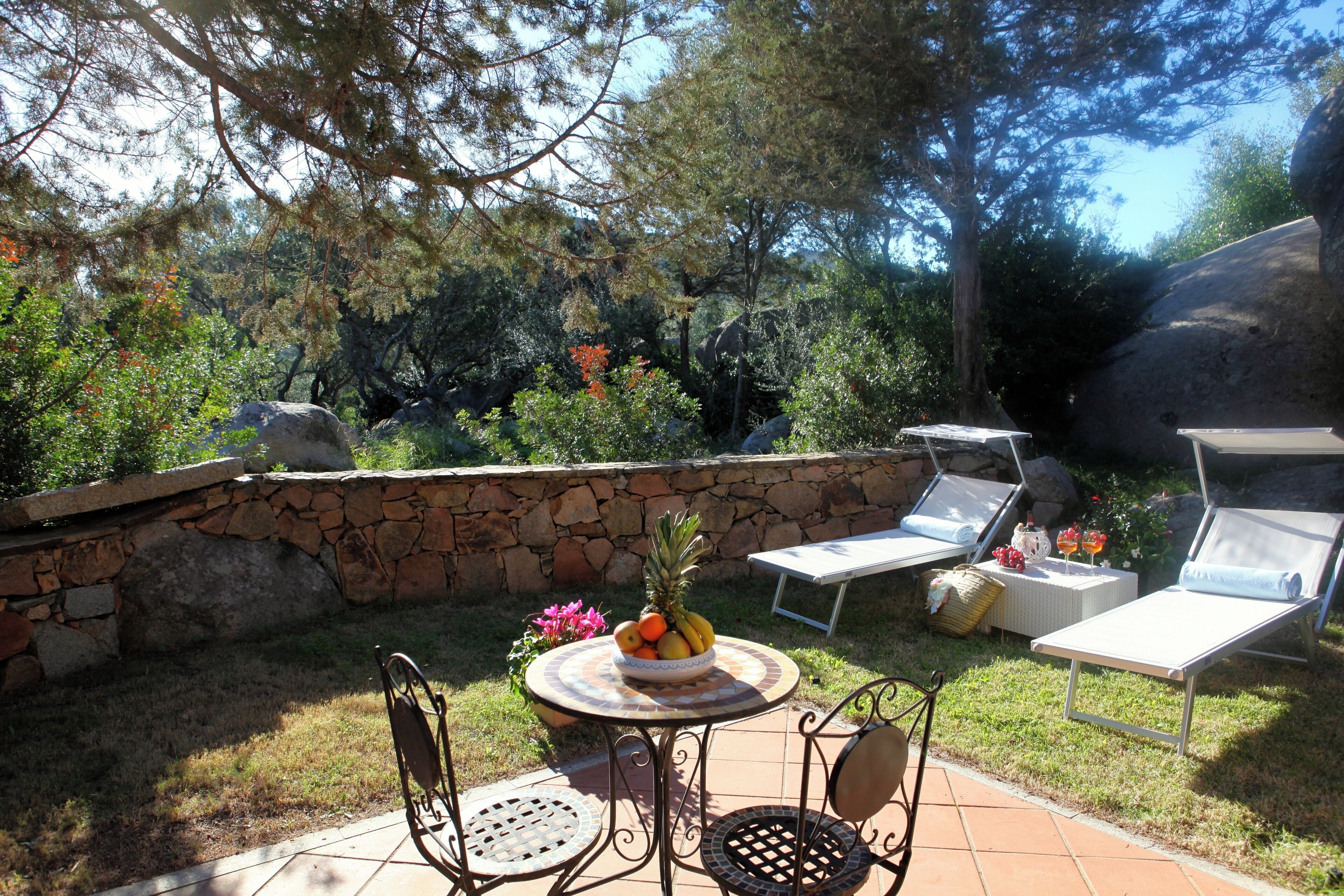 Une terrasse extérieure avec un mur en pierre, une table ronde sur laquelle est posé un panier de fruits, deux chaises longues et une chaise en métal.