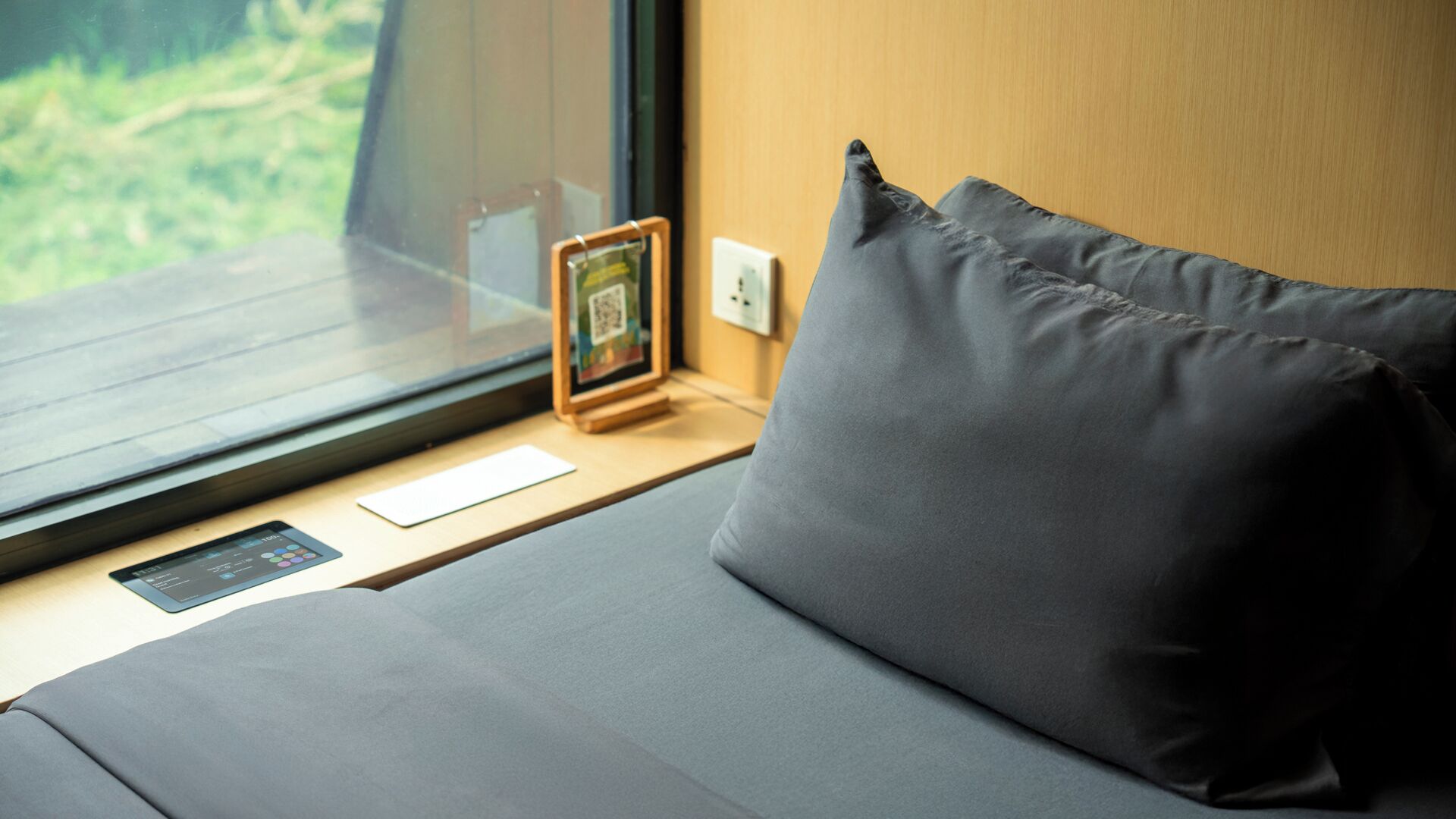 Un lit avec un matelas et des oreillers gris, une petite table sur laquelle se trouvent un smartphone et un cadre photo, et une fenêtre donnant sur un paysage verdoyant.