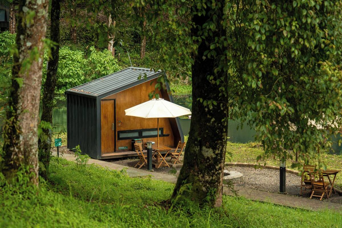 A small wooden cabin with a table and chairs, surrounded by lush greenery and trees.