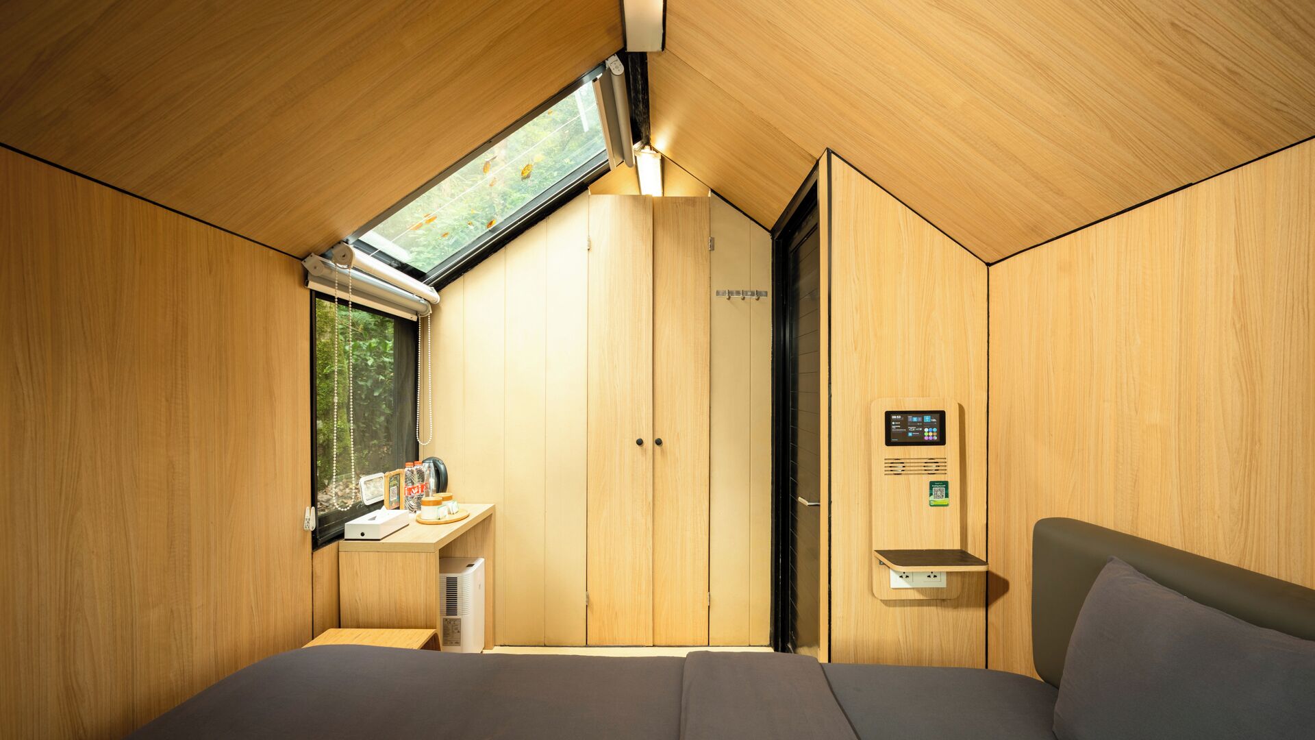 Quarto compacto e moderno com paredes de madeira e teto inclinado, com uma pequena escrivaninha, cama e ar condicionado embutido.