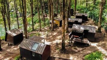 A forested area with several small, dark-colored cabins with sloped roofs, surrounded by trees and a dirt path.
