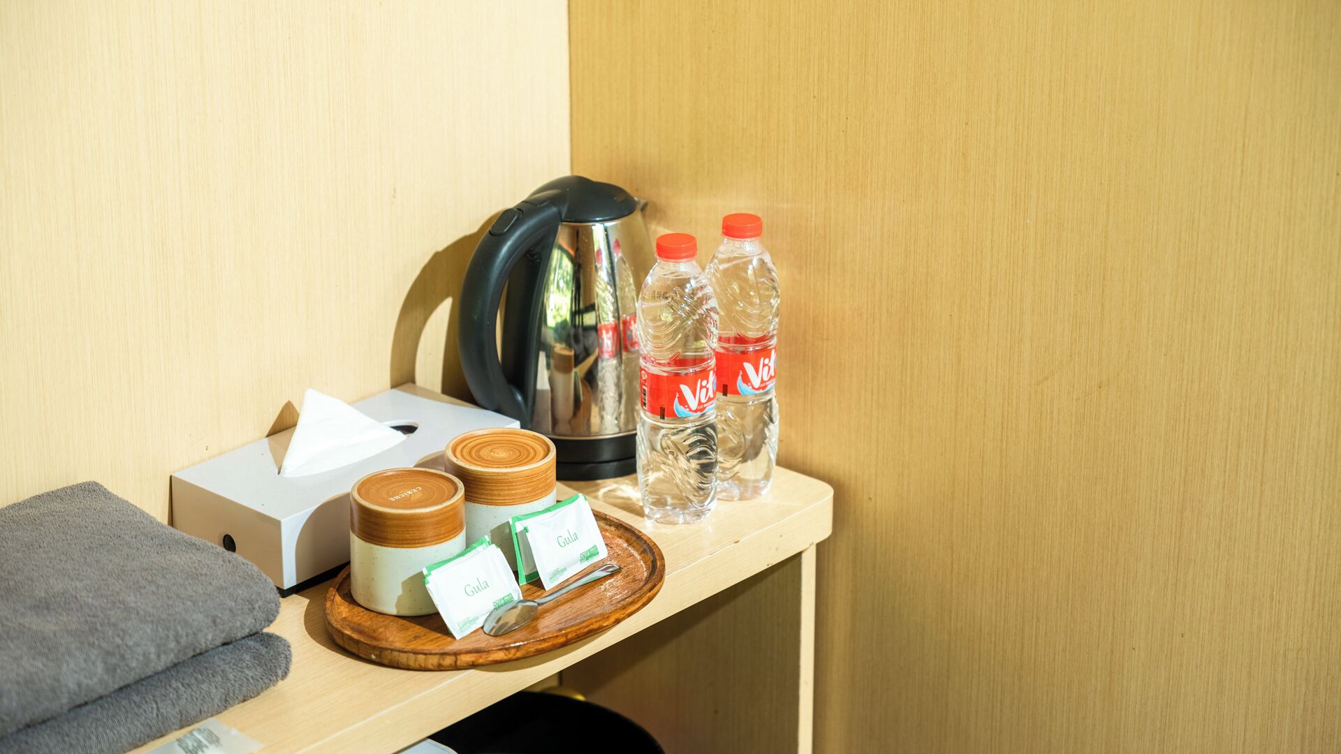 A wooden shelf with a kettle, two bottles of water, a box, and a small container with a spoon.